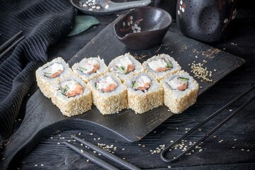 Uramaki inside-out roll with salmon, crab sticks, cream cheese, fresh cucumber and tobiko. Covered with sesame seeds. Black background, black decor.