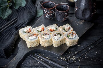 Uramaki inside out roll   with flying fish roe, cream cheese, smoked eel, green onions. Covered with sesame seeds. Black background, black decor.