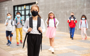 Confident teen girl in protective mask walking outside school building on spring day, going to...