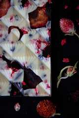 Close-up of a piece of white and dark chocolate bar with raspberry and strawberry pieces on a dark background with flowers of small dried roses