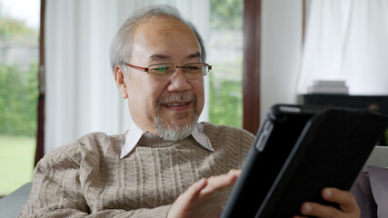 Candid of old asian retired man using tablet computer to transfer money online or financial payment and read text, smile or laugh with happy and positive. Senior asian with digital technology concept