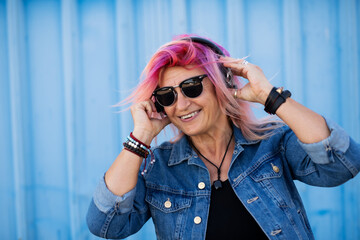 Senior woman with purple pink hair and sunglasses standing against blue background.