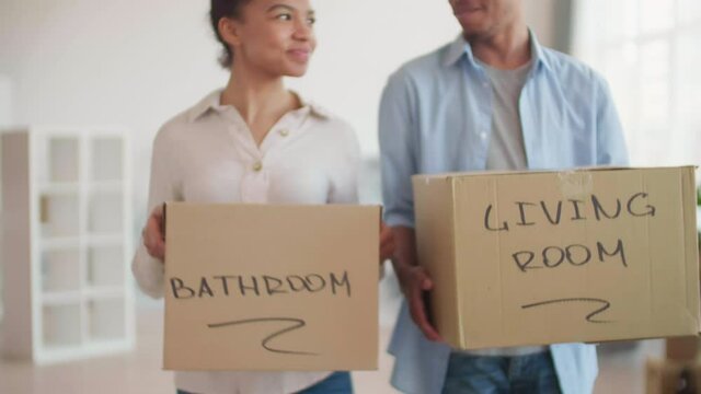 Medium shot of young African-American man and his pretty girlfriend wearing casual clothes going along room with cardboard boxes in hands and communicating