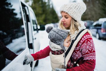 Mother with sleeping small daughter in carrier standing by car in winter nature.