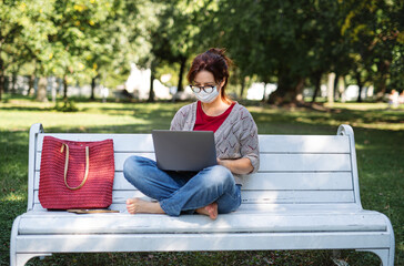 Mature woman working with laptop outdoors in city or town park, coronavirus concept.