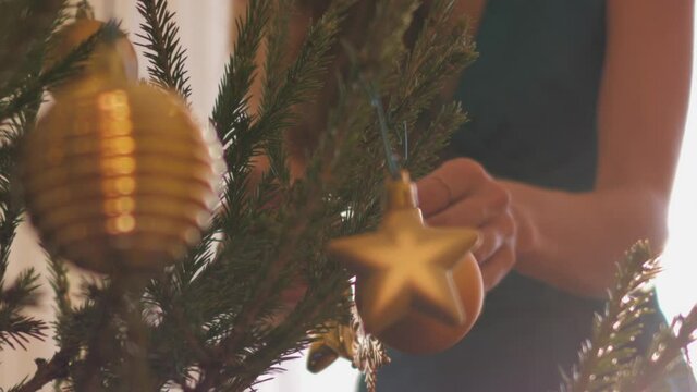 Slim woman with long wavy blonde hair decorates Christmas fir-tree with golden decorations for New Year at home. Concept holiday