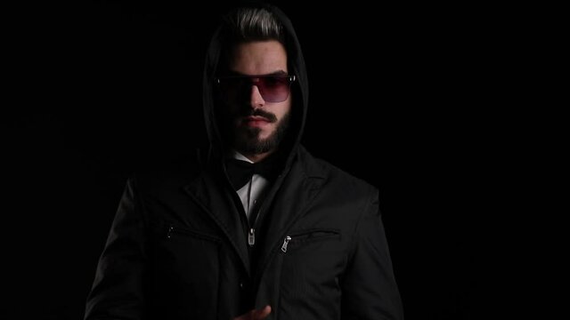 cool young man wearing sunglasses, arranging black jacket, holding hands in pockets and posing on black background in studio