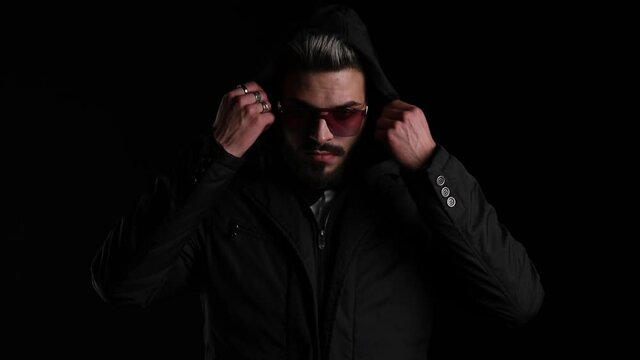 mysterious young man pulling down hoodie, fixing hair, confidently adjusting black jacket and posing on black background in studio