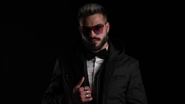 sexy young guy wearing sunglasses, unzipping jacket, holding hands in pockets and looking to side, moving in a side view position and posing on black background in studio