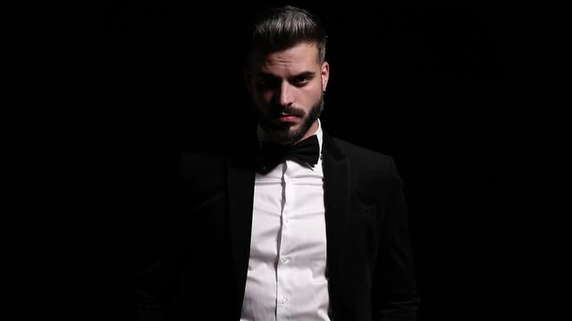 sexy young businessman unbuttoning black tuxedo, fixing bowtie, crossing arms and posing, turning and leaving on black background in studio