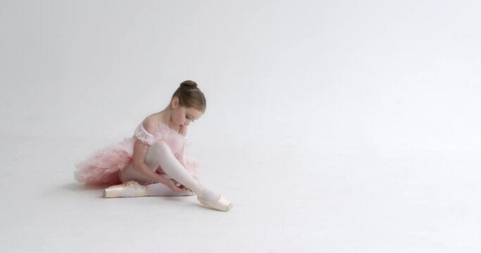 Little girl in a white tutu sits on the floor and ties the ribbons of her Pointe shoes, young ballerina preparing for training, white background.