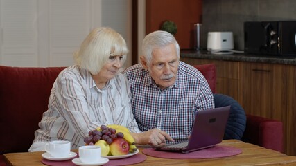 Happy senior elderly couple grandfather and grandmother looking on digital laptop making online shopping. Mature old husband and wife watching news videos, using social media at home