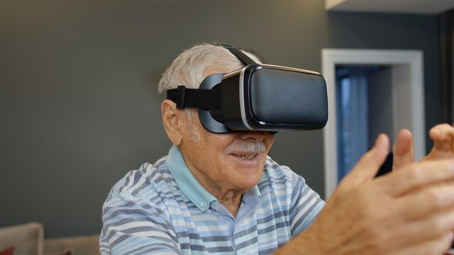 Amazed senior grandfather in virtual headset glasses watching amazing 3D video in 360 vr helmet at home. Future technology. Man in VR goggles playing games, looking around, shows emotion of surprise