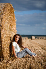 Naklejka na ściany i meble Beautiful young woman near a sheaf of hay in a field. Holidays in the village, a girl enjoying nature in a mown field on a sunny day