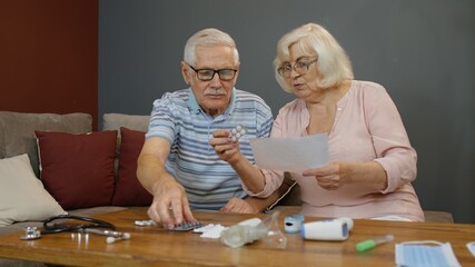 Worried old mature retired grandparents couple looking at medicine prescription, pills, tablets in blisters on table at home. Senior grandfathe, grandmother family on coronavirus quarantine lockdown