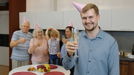 Family celebrating birthday. Portrait of positive handsome man holding glass of champagne. Happy father with wife, daughter and grandparents rejoicing holiday party at home kitchen. Slow motion