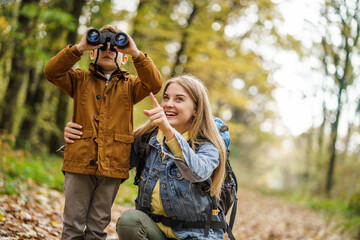 Happy mother and son are hiking in forest. Boy is watching nature with binoculars.