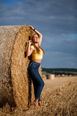 Naklejka na ściany i meble A young beautiful blonde stands on a mown wheat field near a huge sheaf of hay, enjoying nature. Nature in the village