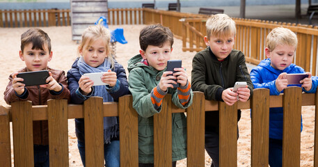 Interested kids playing with smartphones while standing near wooden fence on playground