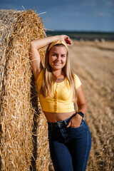 Naklejka na ściany i meble A young beautiful girl in a field stands near a sheaf. Vacation in the village. Young woman in yellow top and jeans