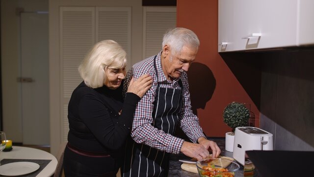 Side View Of Happy Mature Couple In Love Making Dinner. Elderly Woman Hugging From Back Her Husband Cooking Fresh Salad Standing Near Cutting Board In Kitchen At Home. Senior Grandmother, Grandfather