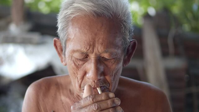 White-haired Asian elderly man do not wear clothes because of hot weather, poor status, more than seventy is sitting smoking tobacco wrapped in nypa fruticans (nipa palm)leaves.