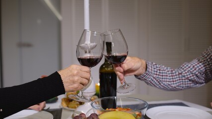 Close up of clinking red wine glasses during romantic dinner. Happy cheerful senior elderly couple dining together in the cozy kitchen, enjoying the meal, celebrating their anniversary