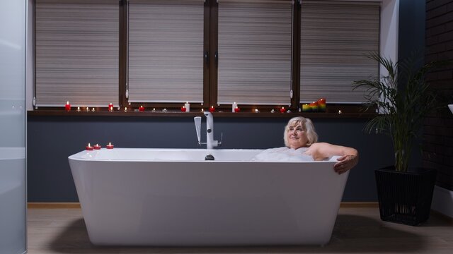 Beautiful Active Senior Woman Lying In Warm Bath With Bubbles, Enjoying Relaxation. Elderly People. Side View Of Cheerful Happy Grandmother At Luxury Home Bathroom In A Romantic Setting With Candles