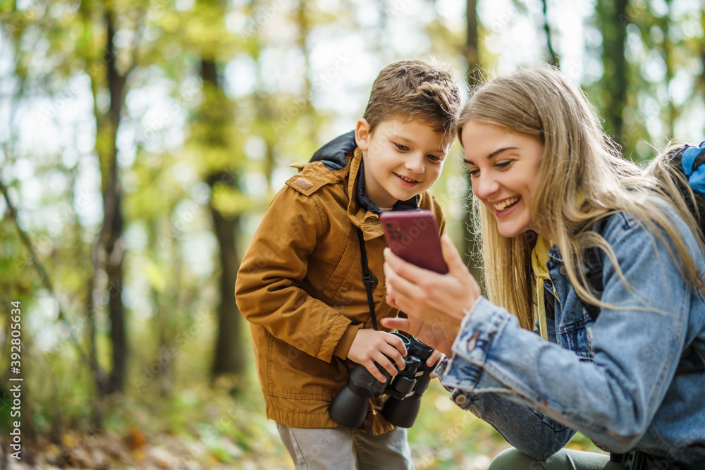 Wall mural happy mother and son are hiking in forest. they are using maps on smart phone.