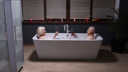 Sexy senior couple grandfather and grandmother is taking foamy bath, drinking red wine in luxury bathroom with candles. Elderly man and woman in love. Spa procedures in hotel. Honeymoon