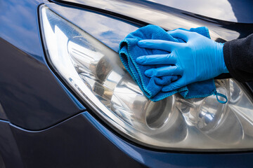 A hand in a blue rubber glove with a blue microfiber rag rubs and polishes the headlight of a car....