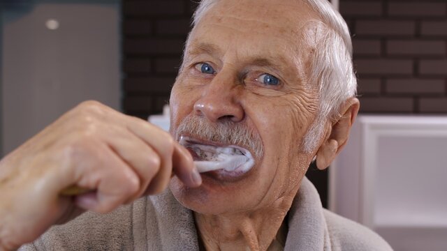 Attractive Old Senior Man Grandfather In Bathrobe Brushing Teeth Looking Into Mirror. Handsome Elderly Grandpa Doing Morning Hygiene After Shower At Luxury Bathroom At Home. Point Of View. POV Shot