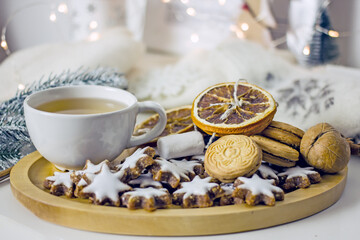 Obraz na płótnie Canvas Homemade christmas tea party background. White mug of tea with asterisk cookies, dried oranges, cinnamon sticks on a white table, surrounded by new decor and garlands. Cozy winter, hygge atmosphere