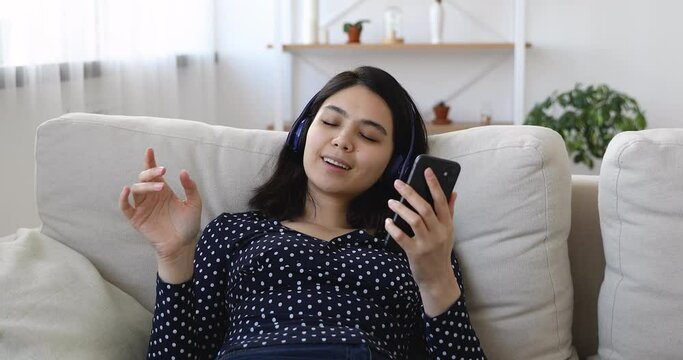Peaceful young asian vietnamese biracial woman relaxing on sofa with closed eyes, listening music from mobile application online library in modern headphones, enjoying hobby leisure pastime at home.