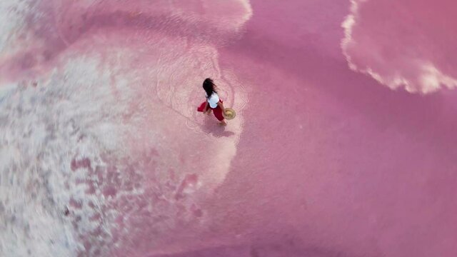 AERIAL, drone follows: happy laughing girl in red skirt runs on shallow water along white salty shore of amazing pink lake with clouds reflections. carefree female showing joy and happiness.