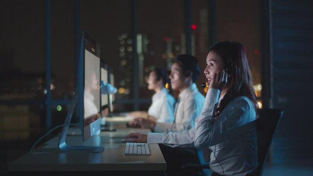 Millennial Asia young call center team or customer support service executive using computer and microphone headset working technical support in late night office. Telemarketing or sales job concept.