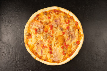 Round hot freshly baked pizza with bacon paprika pineapple and cheese lies on a black stone kitchen table