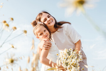 Beautiful young mother and her daughter having fun at the field. Chamomile on foreground, sky background. White clothes.