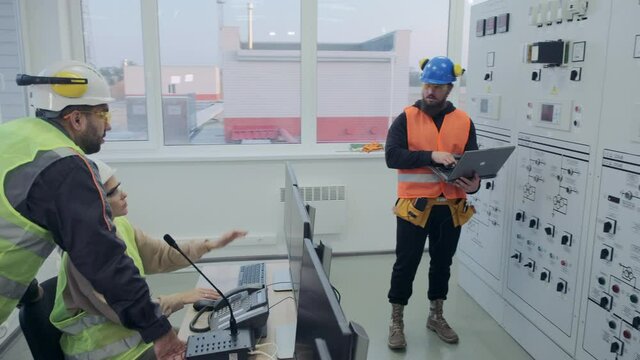 Electrical engineers in helmets sit at computers and brutal colleague observes power station sensors operation on cabinet panel