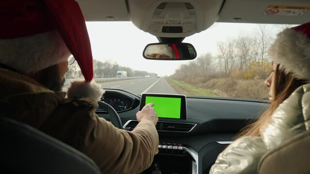 A young couple in Santa hats use a multimedia center with a green screen while sitting in a modern car on the side of the highway. Traveling by car on Christmas Eve