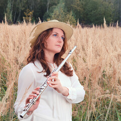 A flute player holds a concert flute while standing in the tall autumn grass. A musician with a wind instrument in the tall grass near the forest © Андрей Журавлев