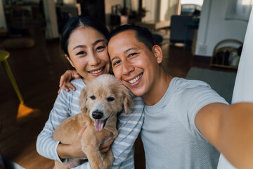 Young adult Asian couple holding a puppy taking a selfie from a phone with home interior in background. 30s mature man and woman with dog pet taking a family photo shots. - Happy group portrait - Powered by Adobe