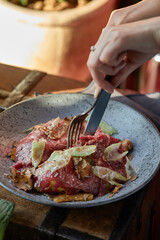 A female hand is preparing a vegetable salad with beef roast beef. A plate of salad with beef roast beef, onions, arugula, pepper and tomatoes on a black table. Top view.