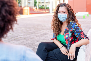 meeting in the open air during a period of coronavirus, social distancing and facial mask, a girl sitting on a bench talks with a friend in front of her con le giuste precauzioni