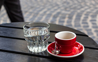 A small red cup with black coffee stands on a red saucer. There is a transparent glass of water nearby. A dark wooden table is located near a small cafe on a narrow street. - Powered by Adobe