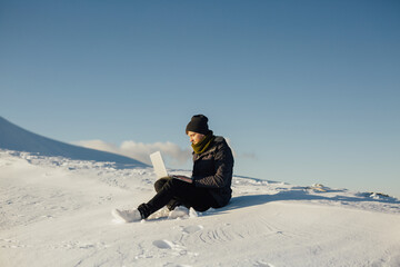 Fototapeta na wymiar Tourist man on the snowy mountain using laptop on mountain peak with blue sky on the background. Copy space.