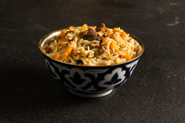 Pilaf is a traditional rice dish with lamb or beef and vegetables in ethnic Uzbek ceramic dishes on a black table.