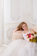 Beautiful young woman with flowers dressed in a white full wide-skirted dress sitting on the sofa