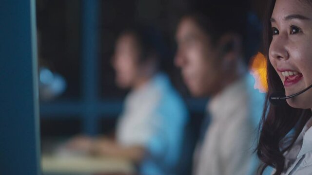 Millennial Asia young call center team or customer support service executive using computer and microphone headset working technical support in late night office. Telemarketing or sales job concept.
