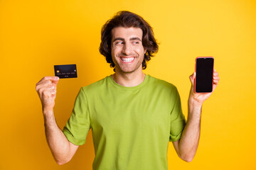 Photo of cheerful man hold phone look screen empty space credit card wear green t-shirt isolated yellow color background